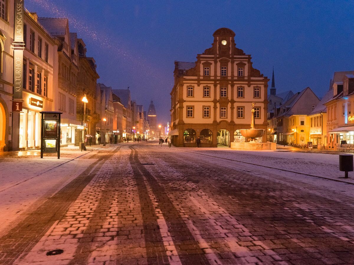 Schneebedeckte historische Altstadtgasse mit beleuchteten Gebäuden, Rathaus und Brunnen am Abend. Wenige Menschen unterwegs.