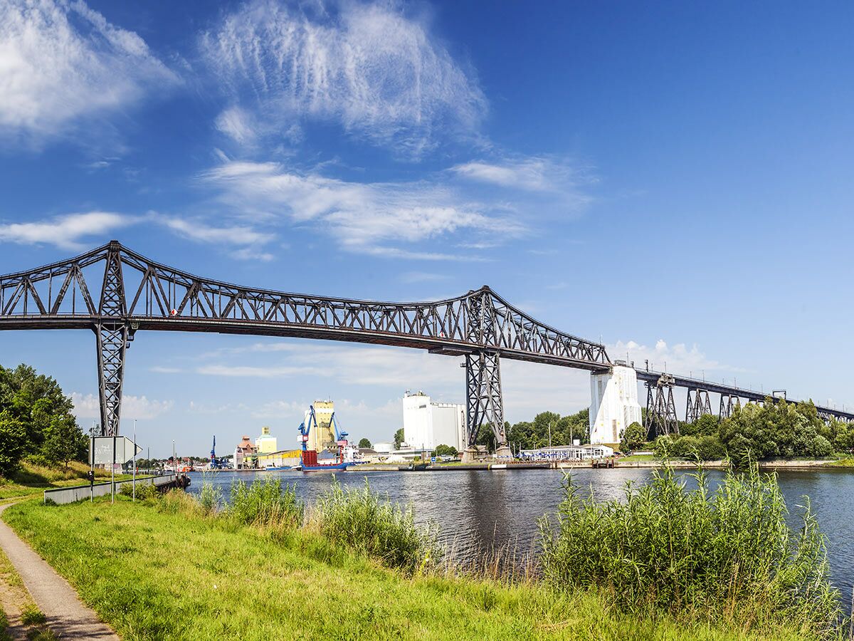 Große Stahlbrücke über den Nord-Ostsee-Kanal mit Industrielandschaft am Ufer und blauem Himmel.