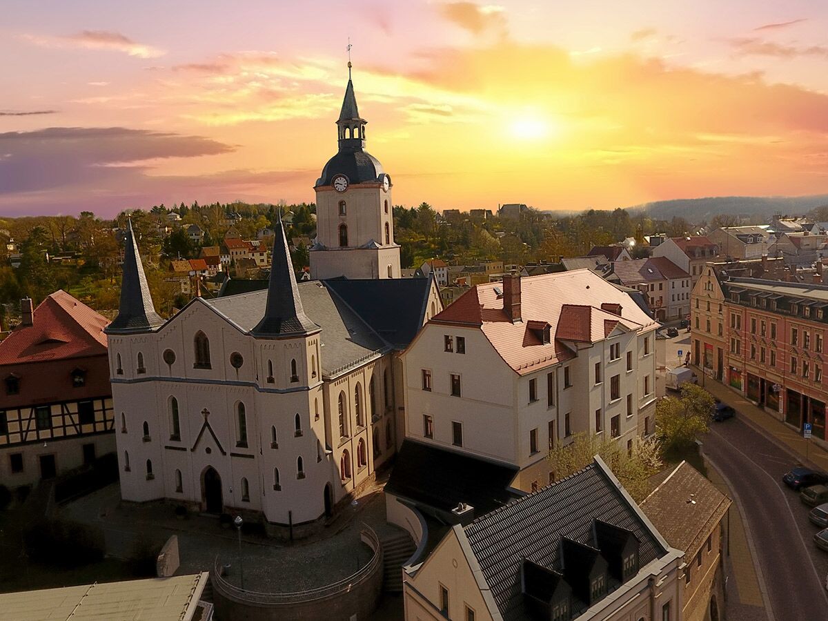 Luftaufnahme einer historischen Stadtkirche mit Doppeltürmen und Glockenturm im warmen Licht des Sonnenuntergangs über einer Stadt.
