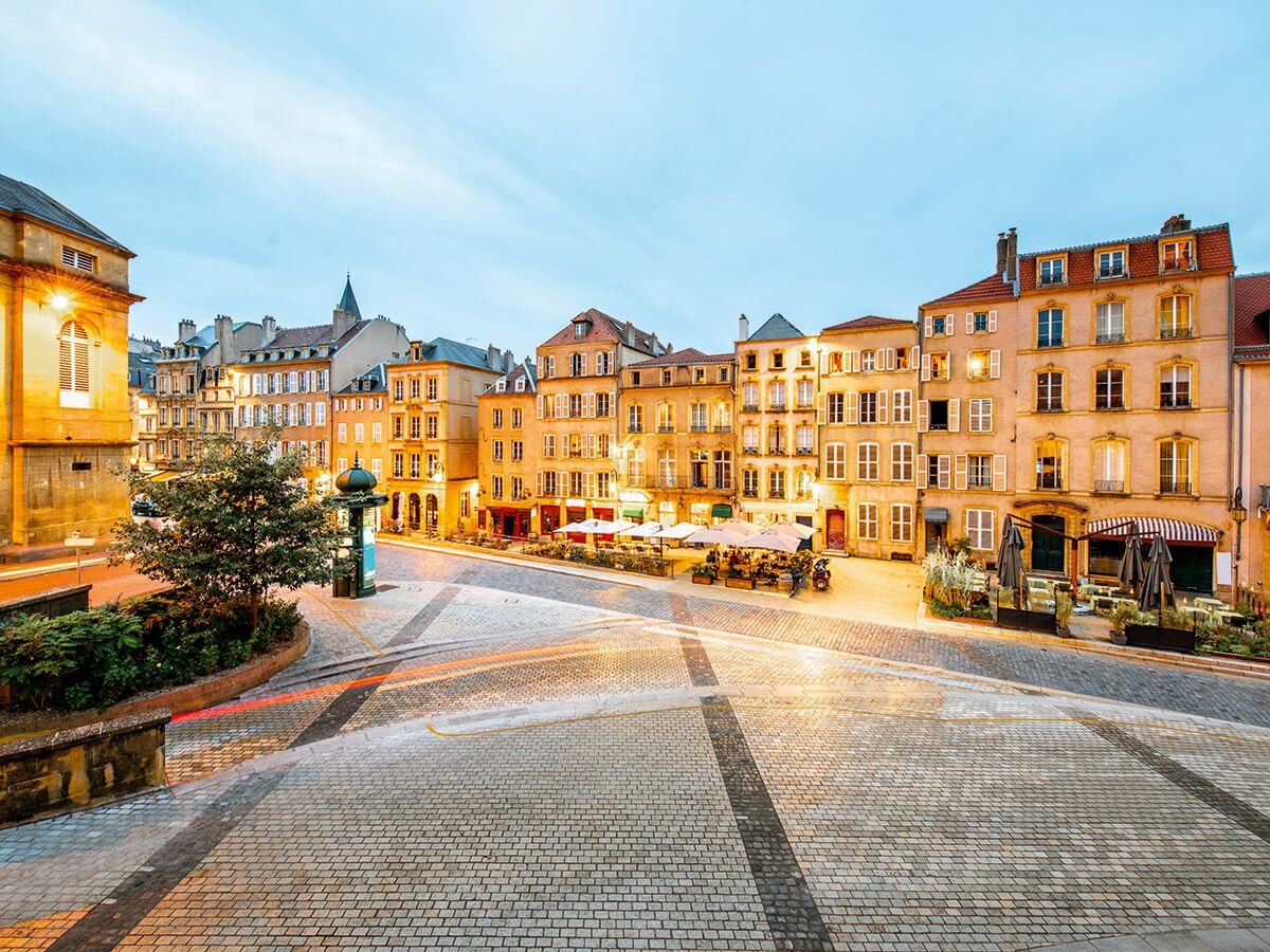 Ein stimmungsvoller historischer Stadtplatz in Metz, Frankreich, bei Dämmerung mit beleuchteten Gebäuden, Cafés und Kopfsteinpflaster.