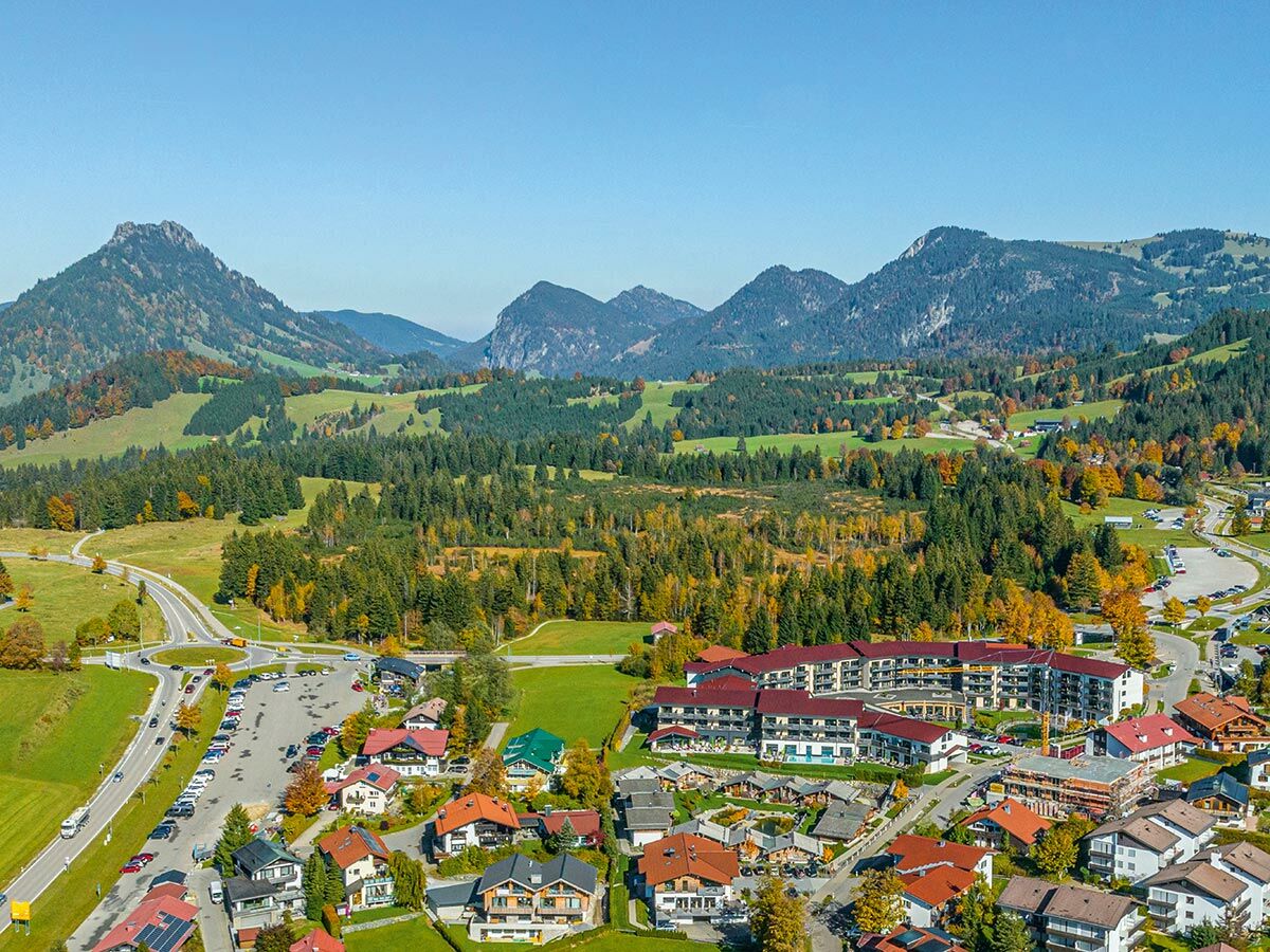 Panoramaaufnahme eines Alpendorfs im Herbst mit bunten Wäldern, Bergen und Häusern. Straße und Parkplatz sind sichtbar.