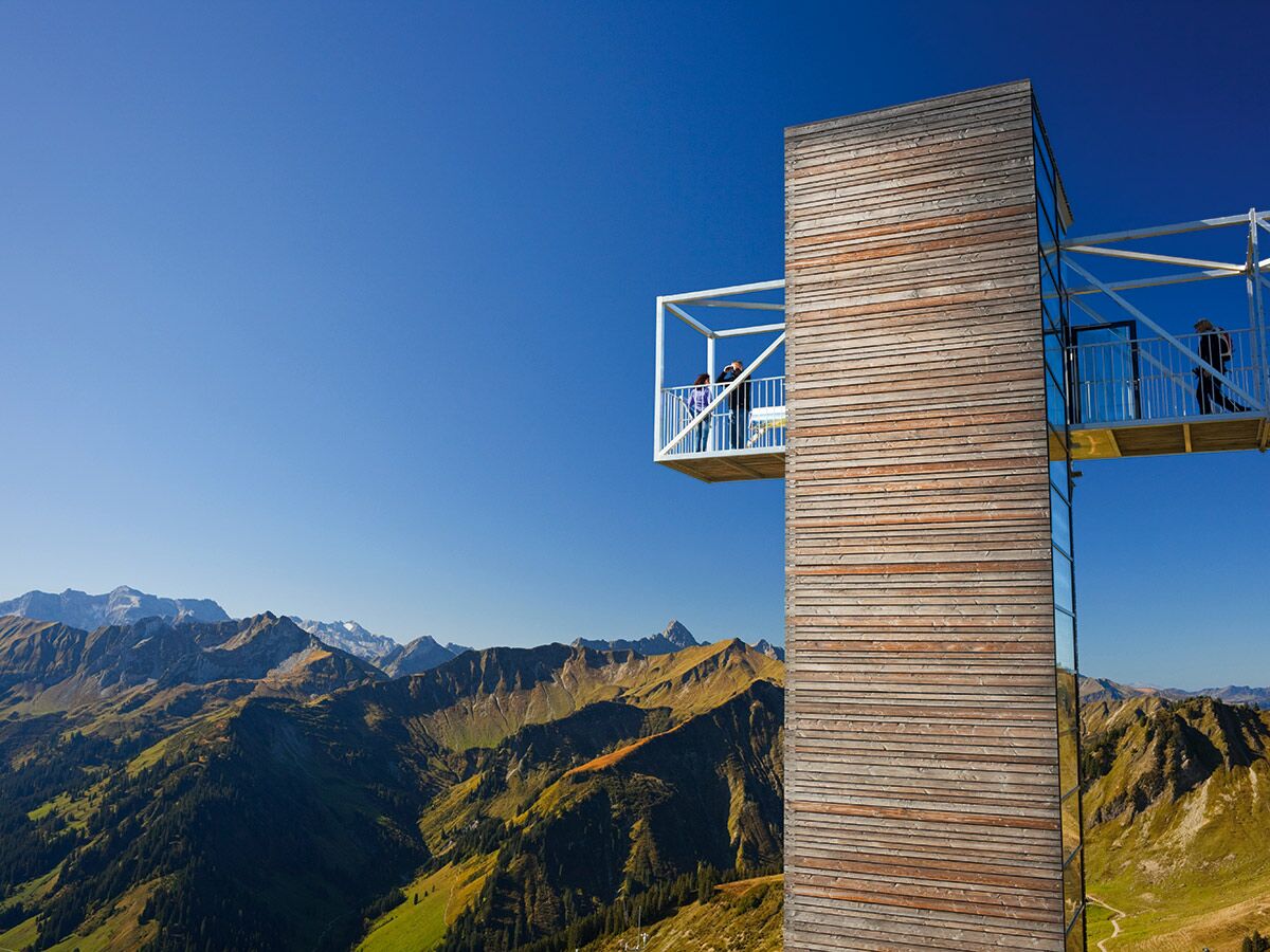 Moderne Holz-Aussichtsplattform über weitem Alpenpanorama mit grünen Tälern, Wäldern und schneebedeckten Gipfeln. Besucher genießen Weitsicht.