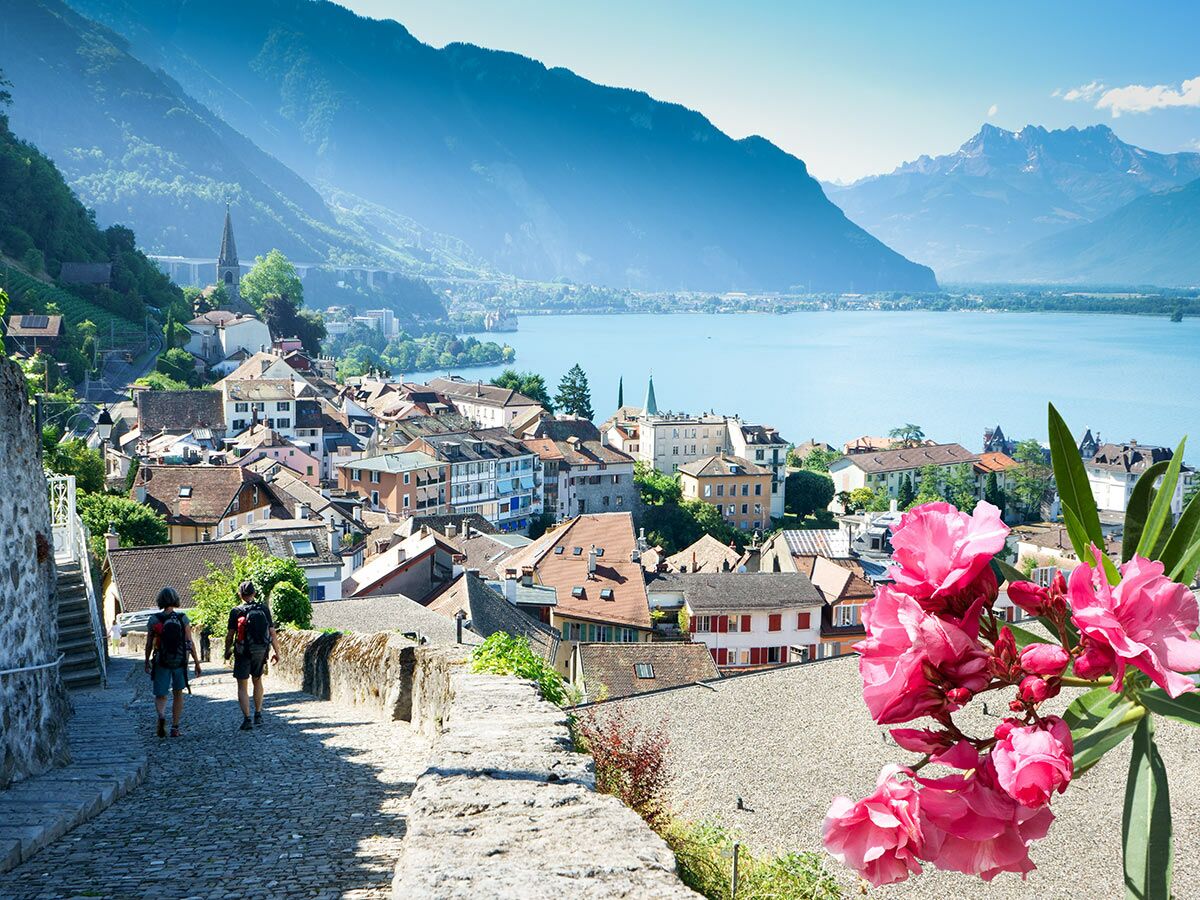 Panoramablick auf die Stadt Montreux am Genfersee mit Bergen, wandernden Menschen und pinken Oleanderblüten.