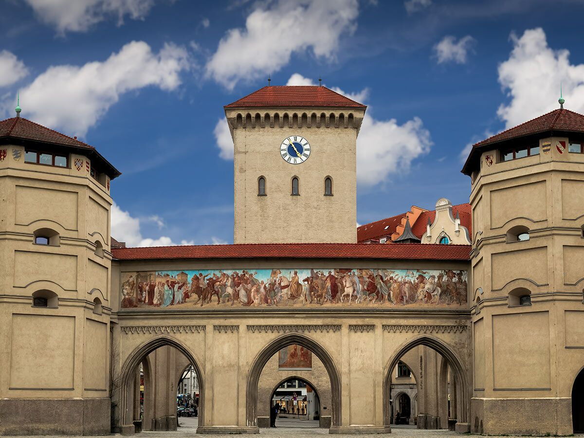 Historisches Sendlinger Tor in München bei blauem Himmel, mit zentralem Turm, Uhr, zwei Seitentürmen und großem Wandgemälde.
