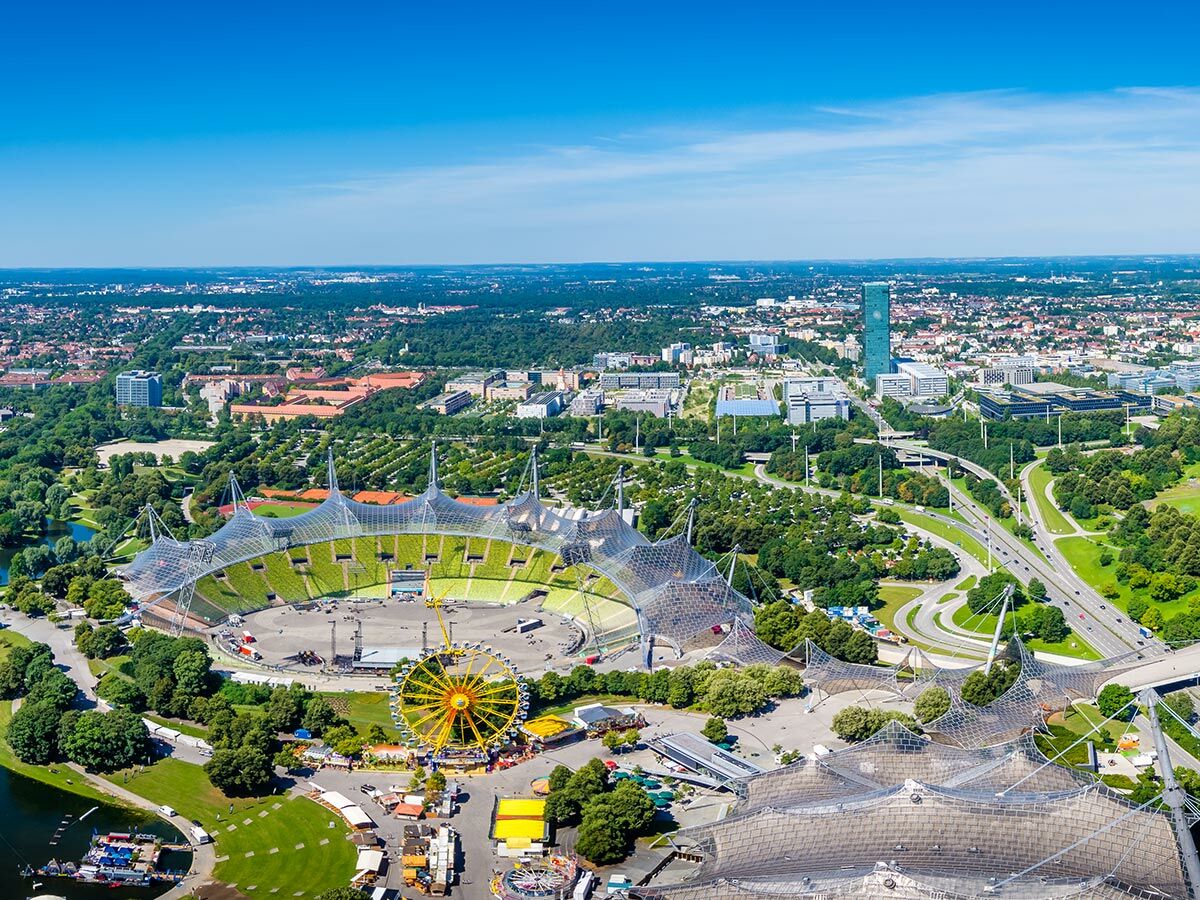 Luftaufnahme des Olympiaparks München mit Stadion, See und Riesenrad unter blauem Himmel, umgeben von der Stadt.