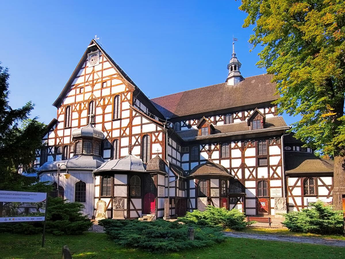 Große historische Fachwerkkirche mit weiß gestrichenen Fassaden und dunklen Holzbalken unter blauem Himmel, umgeben von Grün.