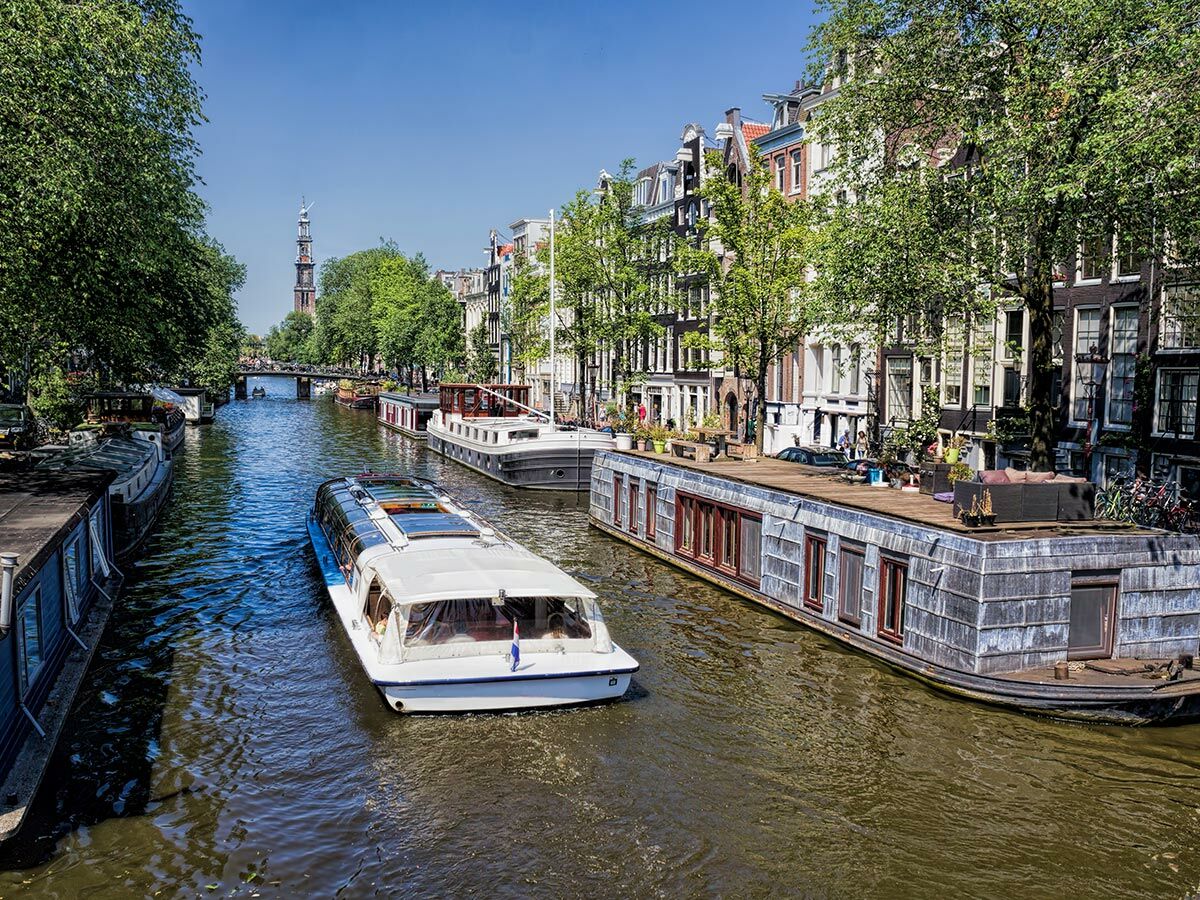 Sonniger Blick auf Amsterdamer Kanal mit Touristenboot, Hausbooten, historischen Grachtenhäusern und Westertoren im Hintergrund.
