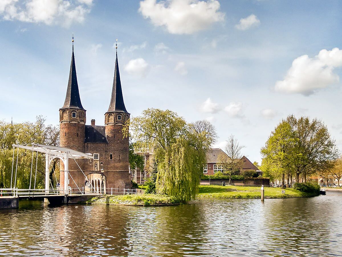 Historische Oostpoort in Delft, Niederlande, mit zwei Türmen, weißer Zugbrücke über einem Kanal und grünen Bäumen.