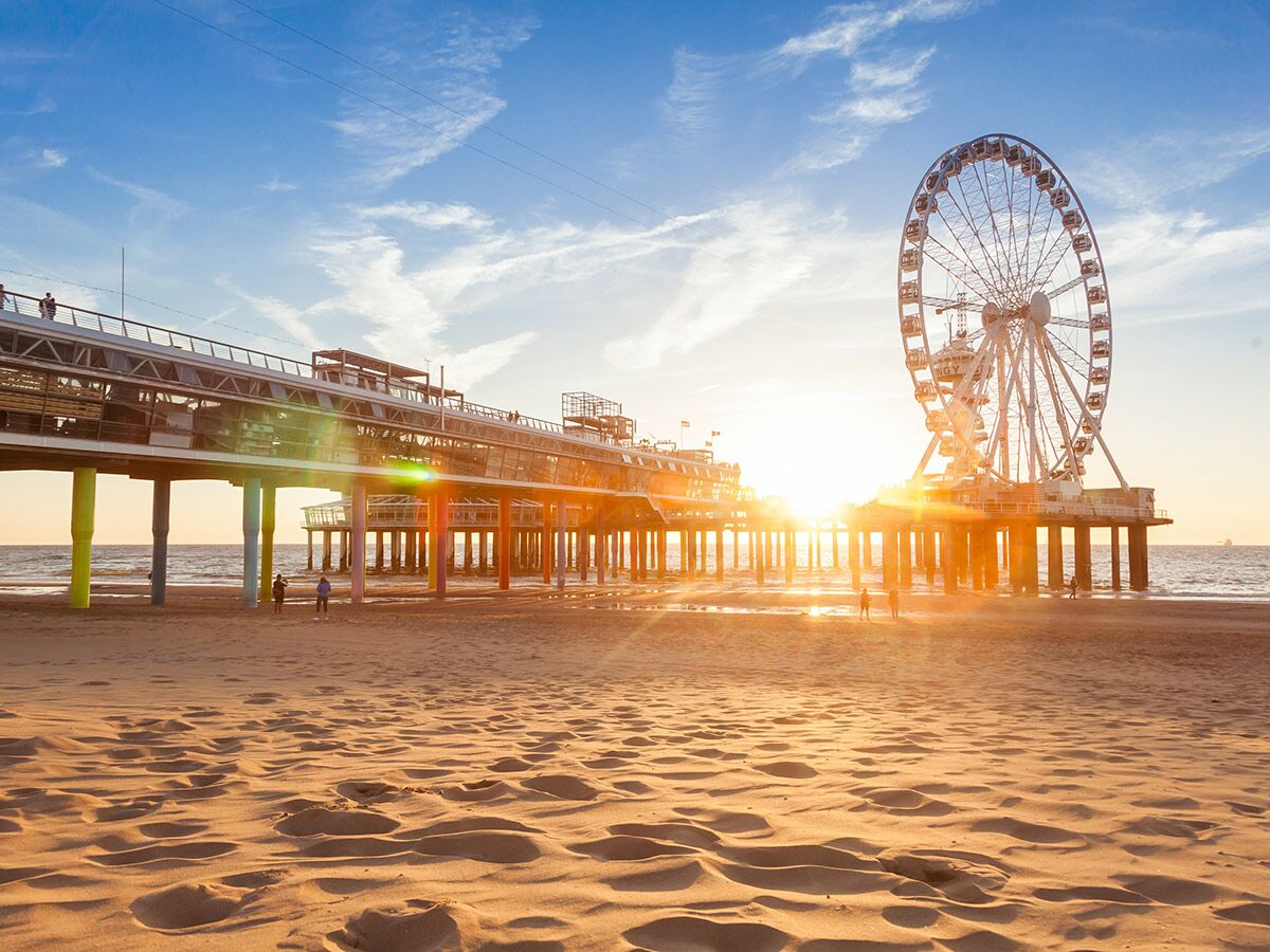 Goldener Sonnenuntergang am Strand mit großem Riesenrad und Pier. Menschen spazieren am Meer entlang bei warmem Licht.