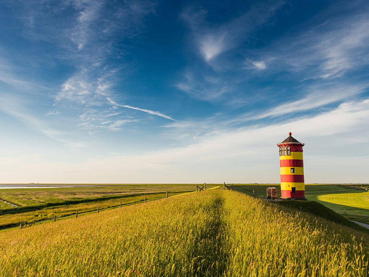 Rot-gelb gestreifter Leuchtturm auf grünem Deich vor weiter Küstenlandschaft mit blauem Himmel.