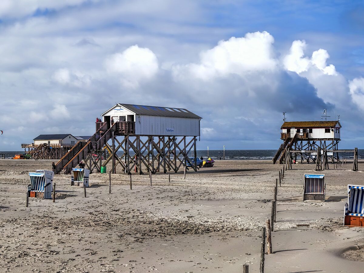 Weite Sandstrandlandschaft mit traditionellen Pfahlbauten, blauen Strandkörben und bewölktem Himmel an der deutschen Nordseeküste.