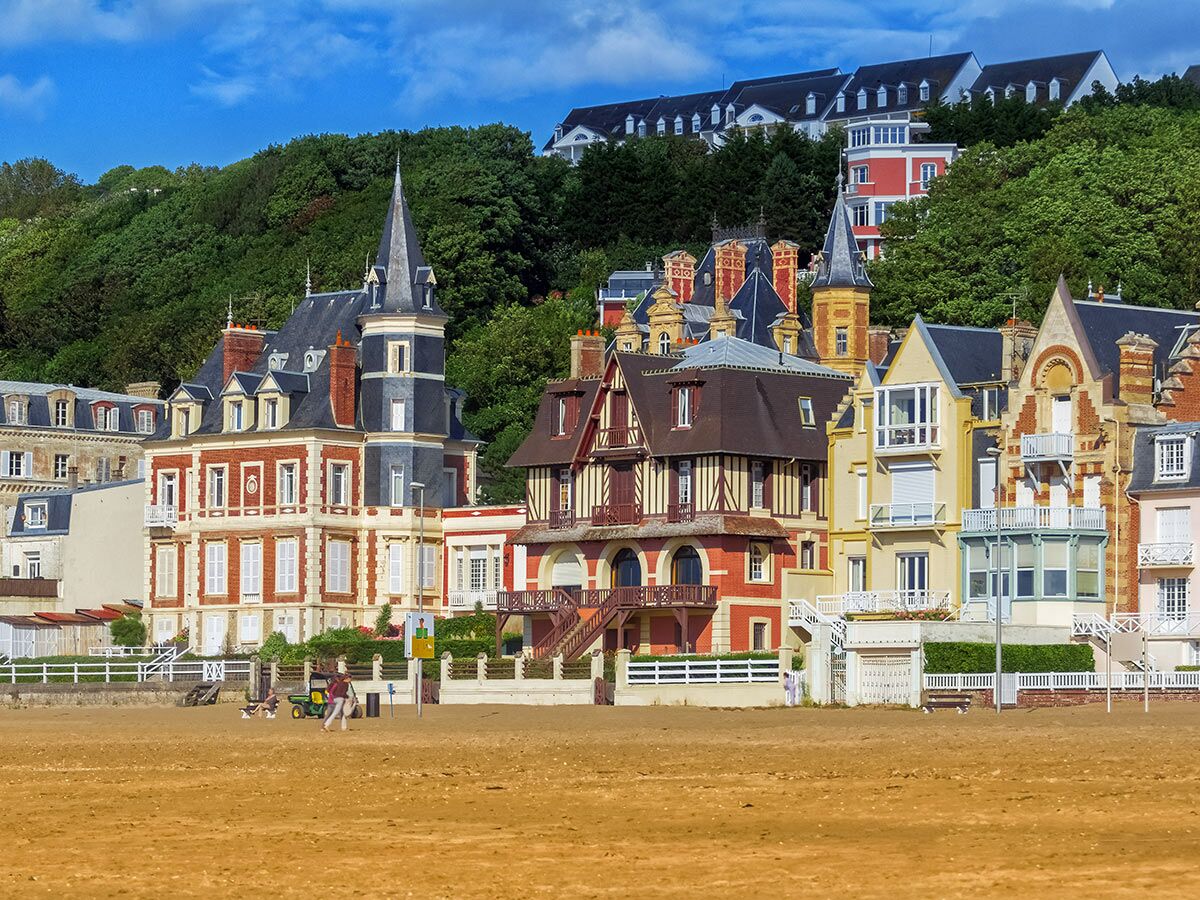 Prächtige historische Villen und bunte Häuser mit Türmen an einem Sandstrand vor grünen Hügeln unter blauem Himmel.