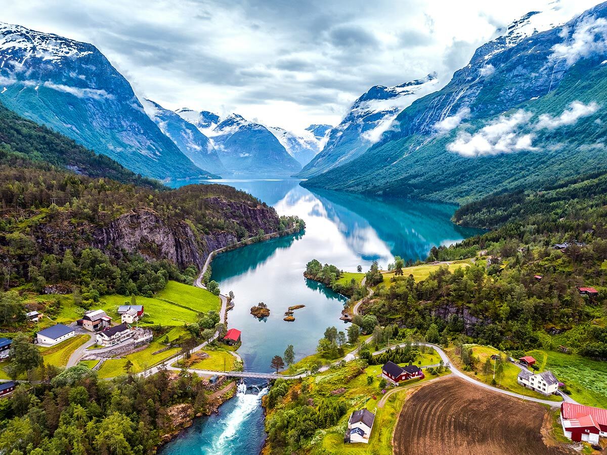 Malerischer norwegischer Fjord mit schneebedeckten Bergen, türkisfarbenem Wasser, üppigen Wäldern und einem kleinen Dorf am Flussufer.