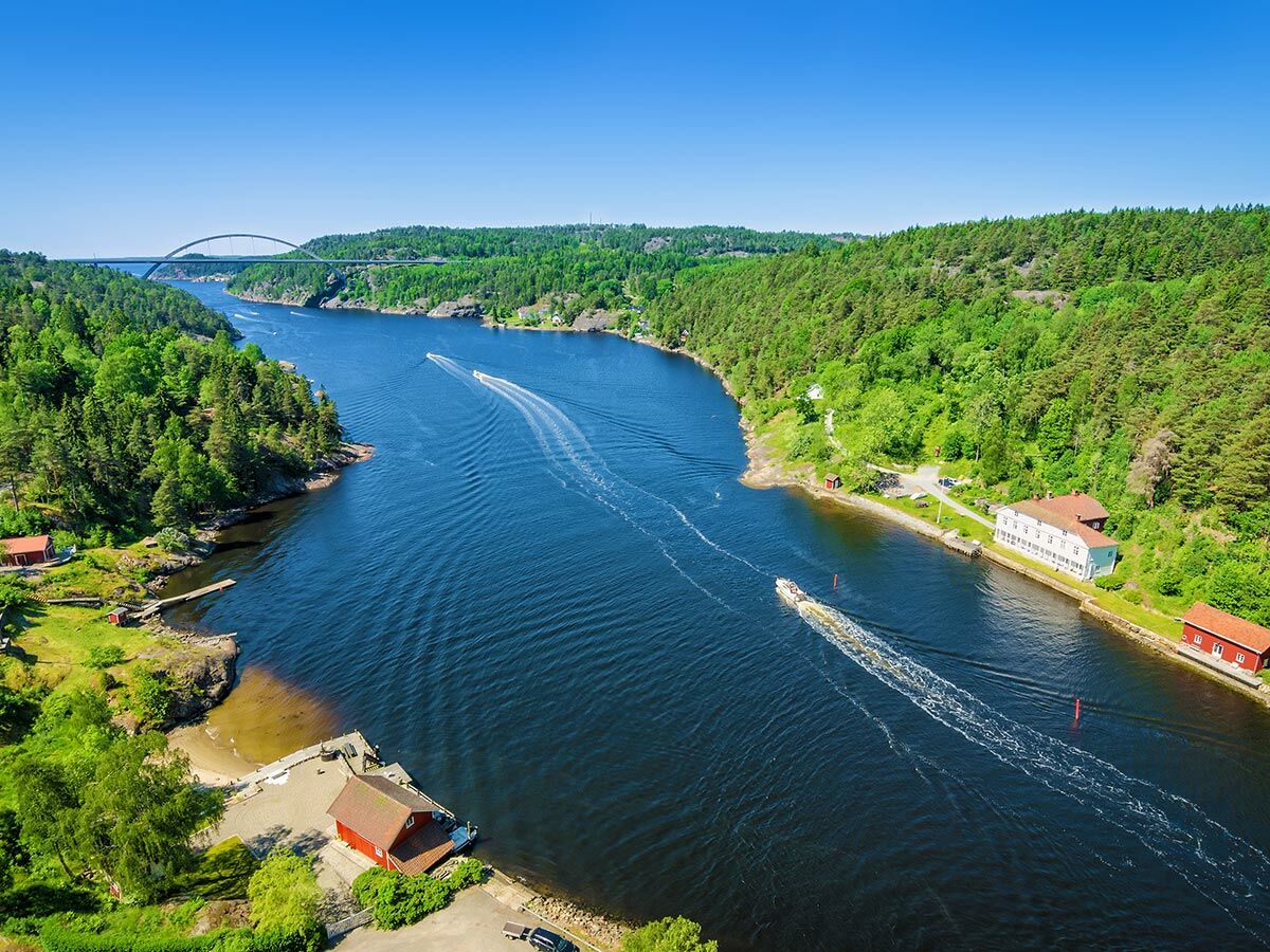 Luftaufnahme einer weiten Flusslandschaft mit grünen Wäldern, einer großen Brücke im Hintergrund und mehreren Booten auf dem Wasser.