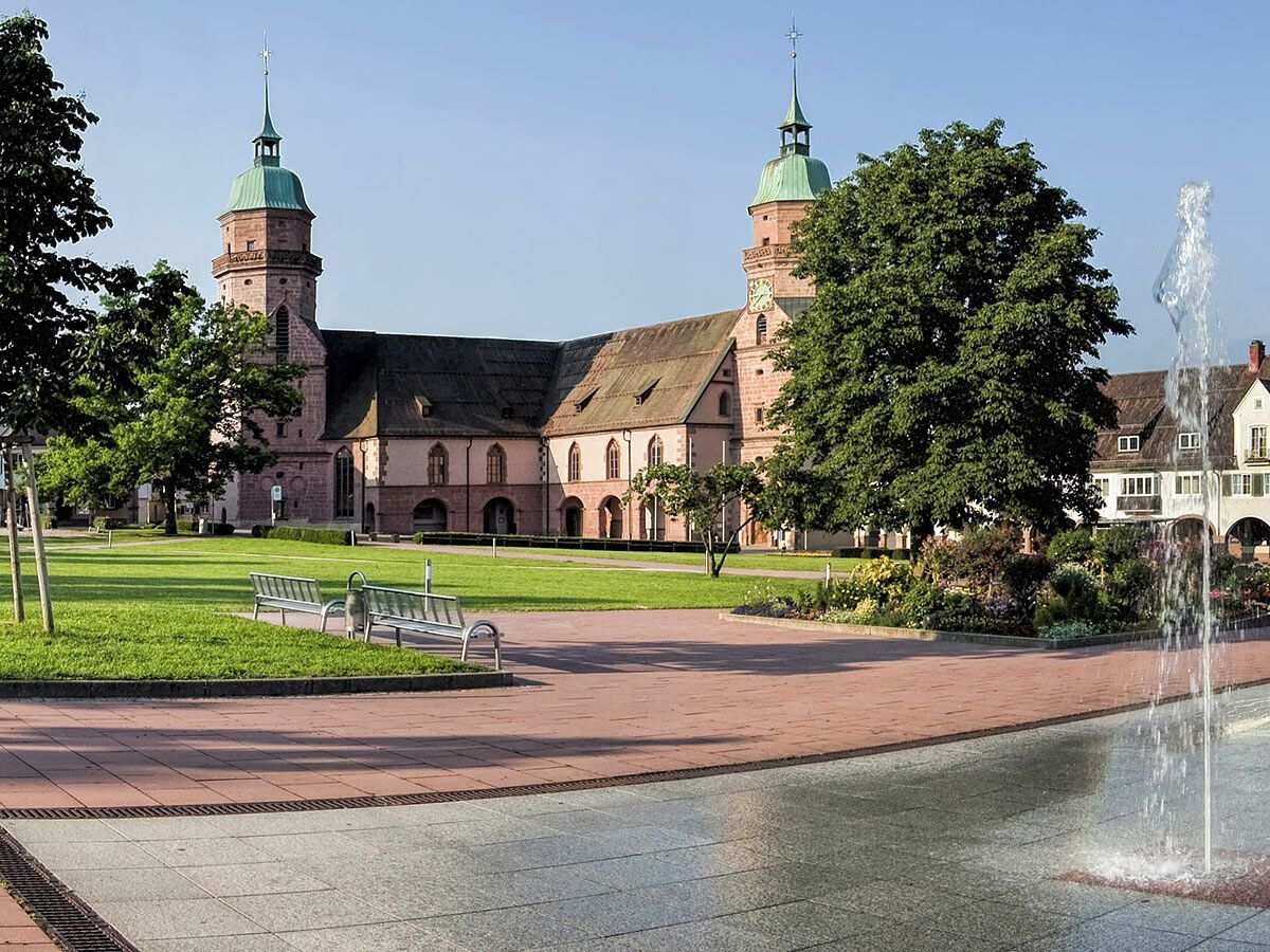 Historische Maulbronner Klosteranlage mit zwei Türmen, Grünflächen und modernem Brunnen bei Sonnenschein.