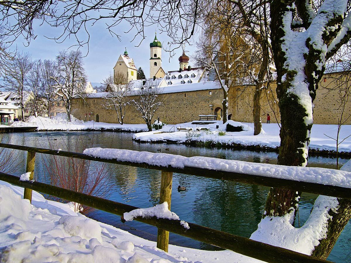 Verschneite historische Stadtmauer und Gebäude am Fluss mit einem schneebedeckten Zaun im Vordergrund und einer Person.