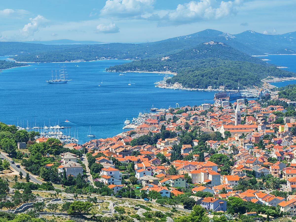 Panorama von Mali Lošinj, Kroatien, mit roter Dächerstadt, blauem Meer, Hafen, Booten, Segelschiff und grünen Bergen.