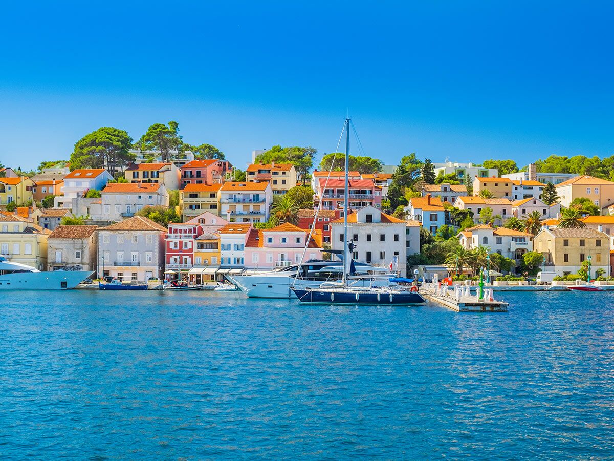 Blick auf eine malerische Küstenstadt mit vielen bunten Häusern und traditioneller Architektur am Meer. Mehrere Yachten liegen im Hafen unter blauem Himmel.