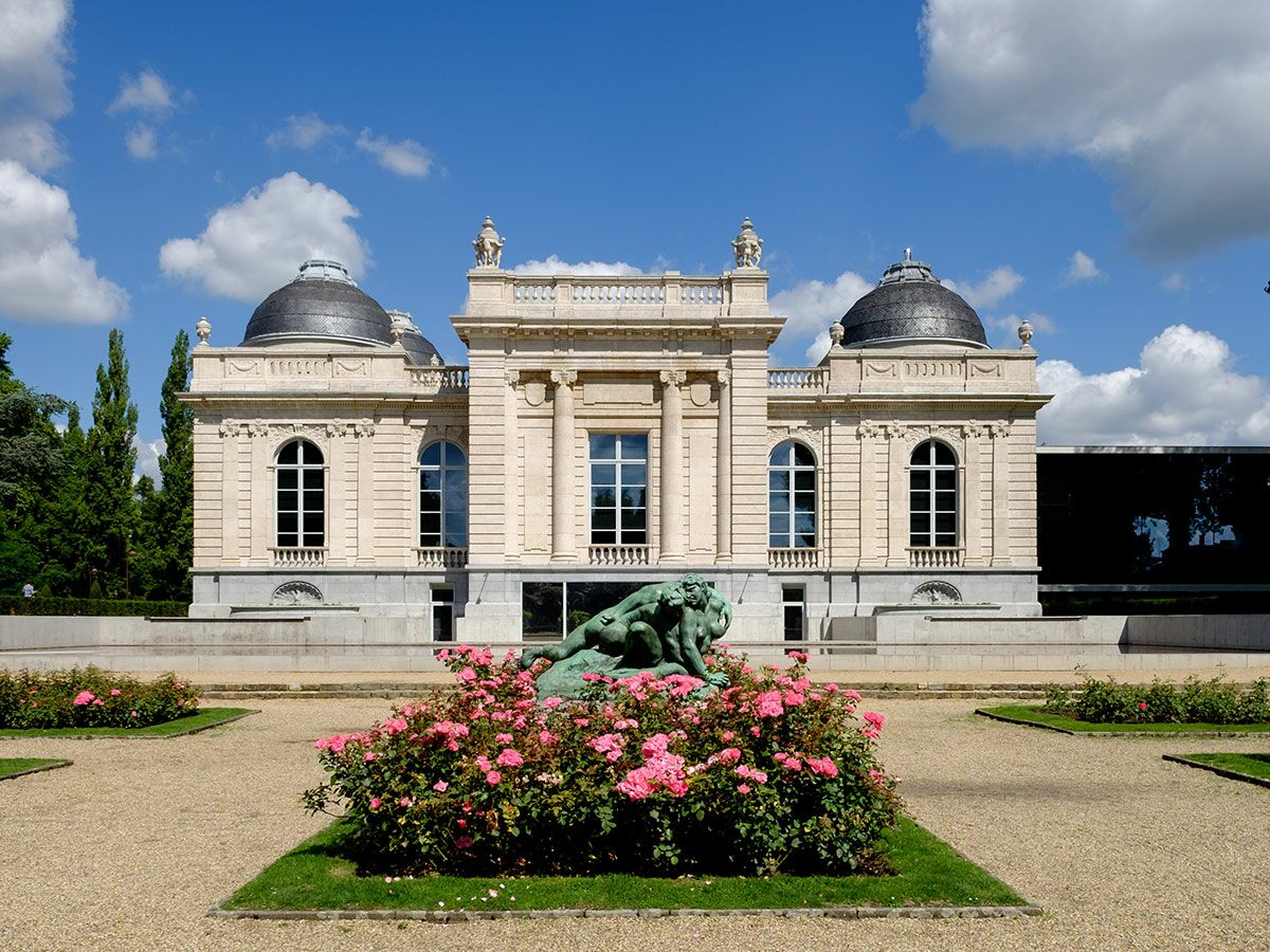 Großes neoklassizistisches Schloss mit zwei Kuppeln, Bronzeskulptur, rosa Rosen und Kiesweg unter blauem Himmel.