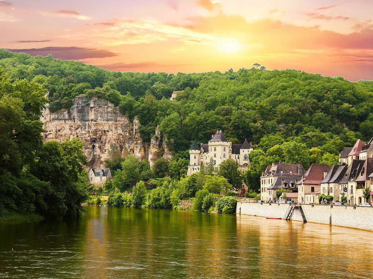 Panoramablick auf ein historisches Dorf am Fluss Dordogne mit majestätischem Schloss, grünen Klippen und goldenem Sonnenuntergang.