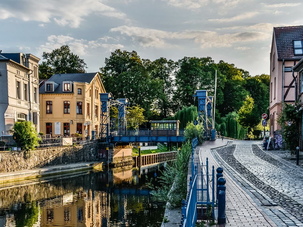 Malerische Altstadtansicht am Kanal mit historischer Zugbrücke, Kopfsteinpflasterstraße und sonnenbeschienenen Gebäuden.