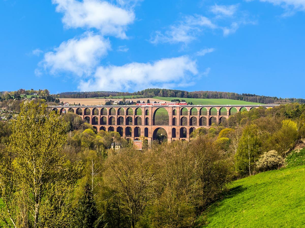 Ein roter Zug überquert ein imposantes, mehrstufiges Ziegelstein-Viadukt in einer grünen Frühlingslandschaft unter blauem Himmel.