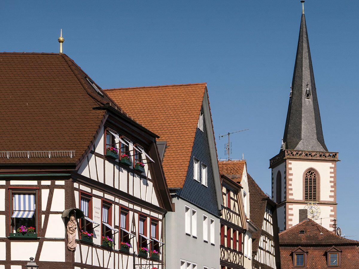 Reihe traditioneller Fachwerkhäuser mit Giebeldächern und Blumentöpfen unter strahlend blauem Himmel, daneben ein Kirchturm mit Uhr.