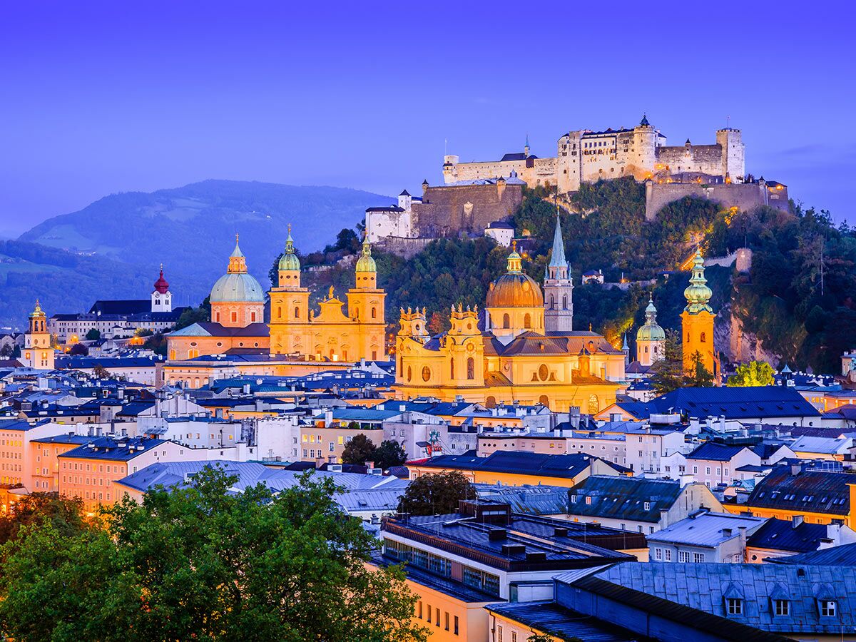 Panoramablick auf Salzburg bei Dämmerung, mit der beleuchteten Festung Hohensalzburg und historischen Kirchen im Vordergrund.