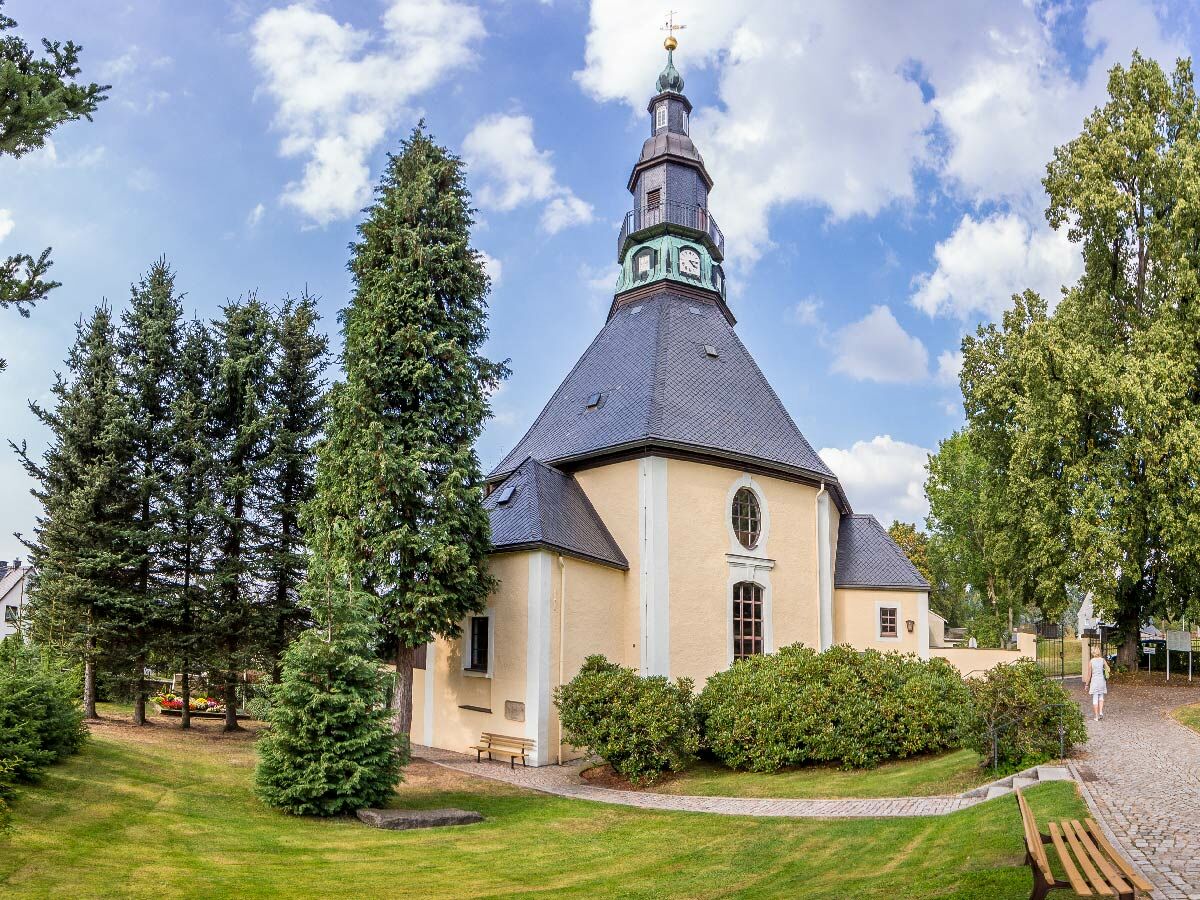 Historische Kirche mit hellem Putz, dunklem Schieferdach und hohem Turm unter blauem Himmel, umgeben von Bäumen und Rasen.