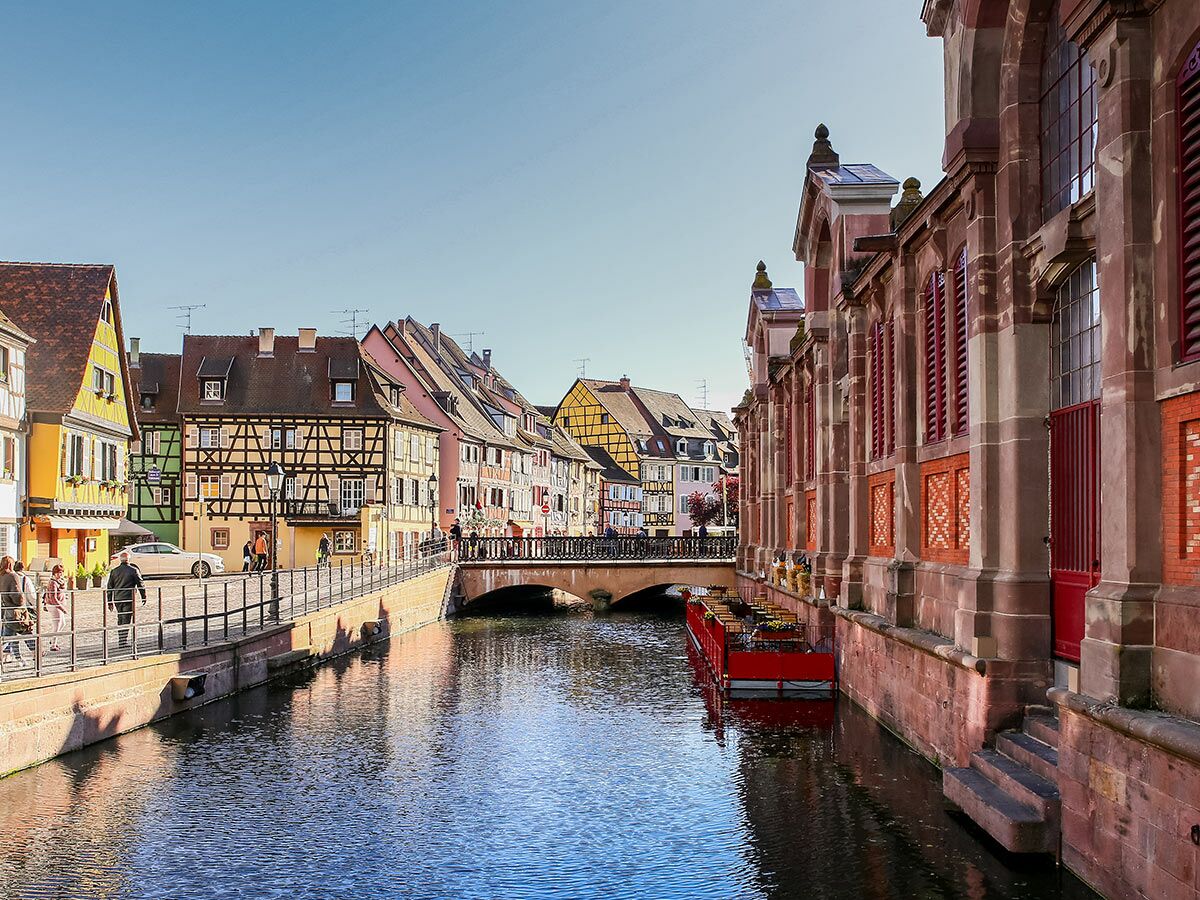 Malerisches Colmar: Kanal, bunte Fachwerkhäuser, historisches Gebäude, Brücke, Menschen und Boot unter blauem Himmel.