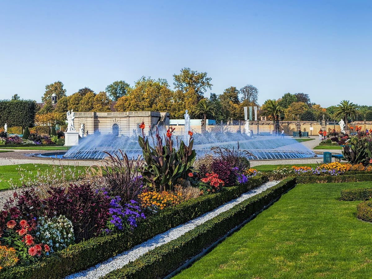 Großer, gepflegter Schlosspark mit einem imposanten Springbrunnen, umgeben von weißen Statuen, bunten Blumenbeeten und grünen Rasenflächen unter blauem Himmel.