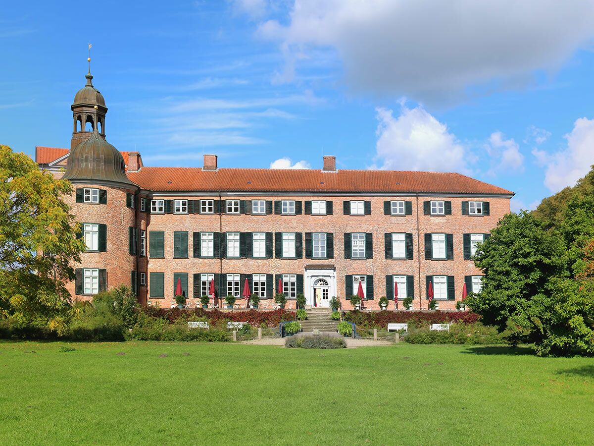 Außenansicht von Gut Billerbeck: historisches Backsteingebäude mit Kupfertürmchen, roten Dächern, grünen Fensterläden und Gartenanlage.