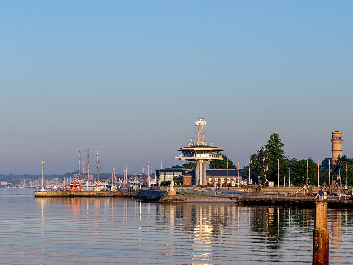 Blick über die ruhige Kieler Förde auf einen modernen Schifffahrtsturm, einen historischen Backsteinturm und zahlreiche Segelboote am Anleger bei Sonnenlicht.