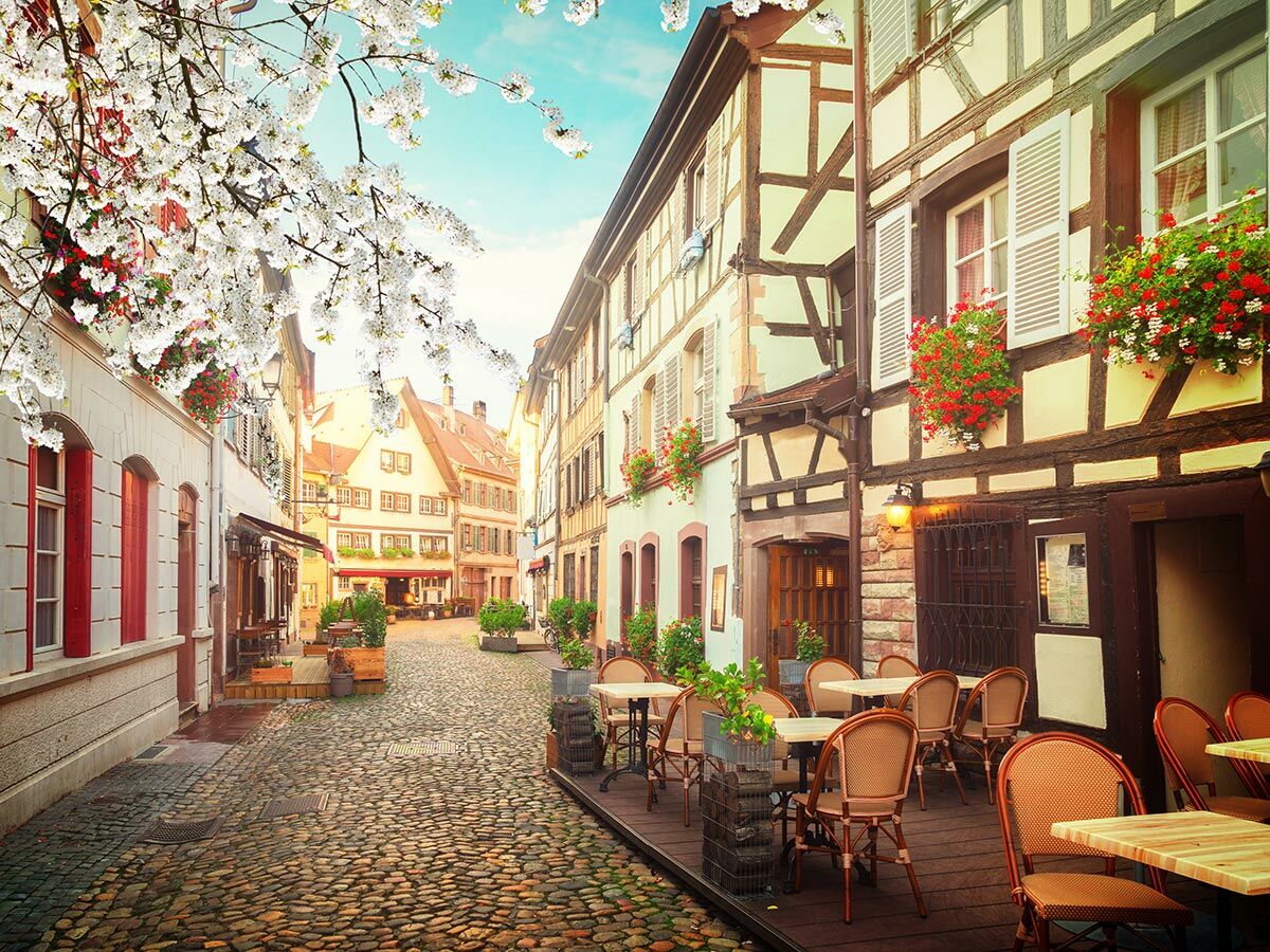 Malerische europäische Gasse mit Fachwerkhäusern, blühenden Kirschbäumen und Café-Tischen unter blauem Himmel am Tag.