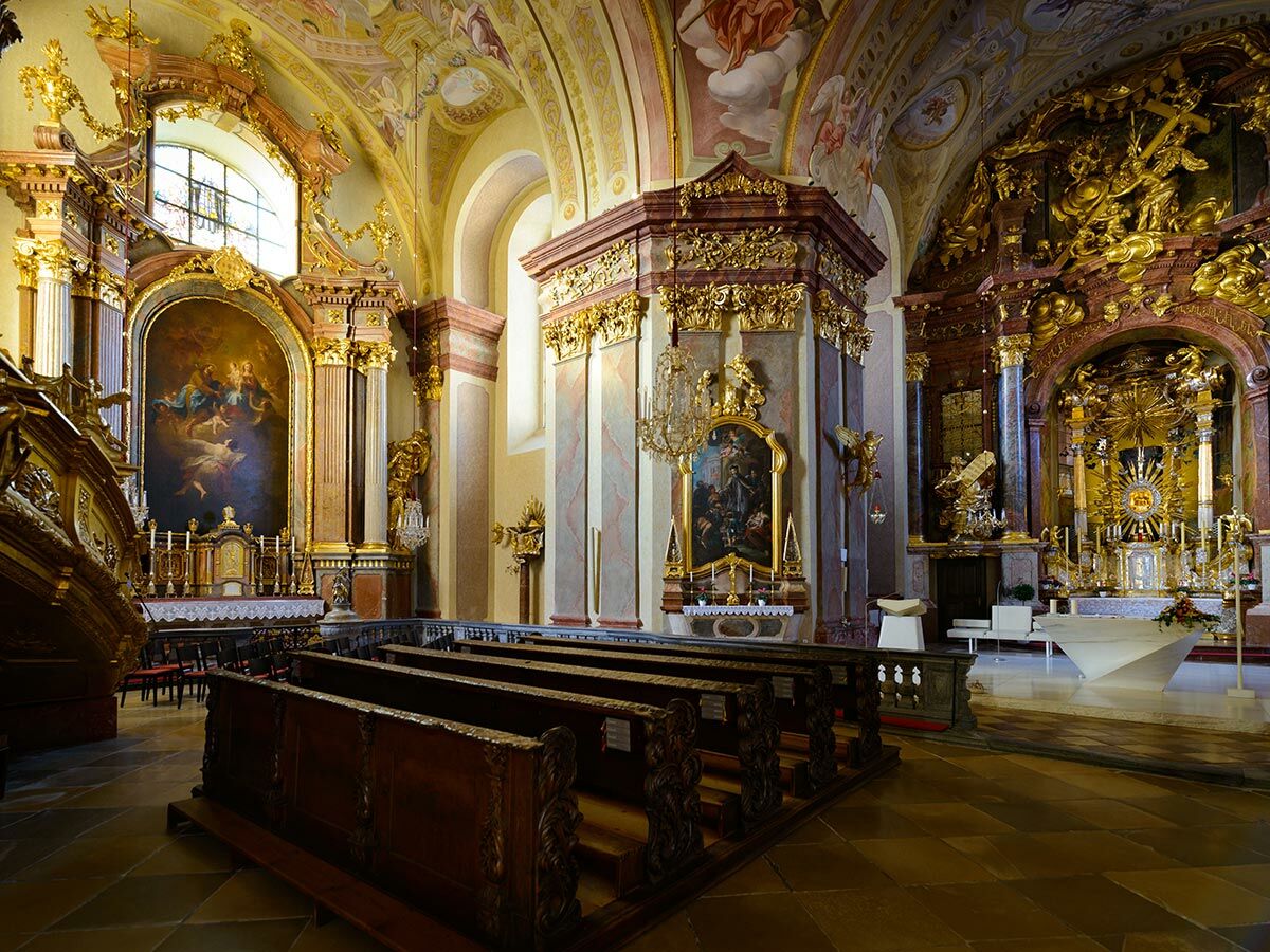 Opulente Innenansicht einer Barockkirche mit goldenen Altären, Deckenfresken, dunklen Kirchenbänken und Marmordekoration.