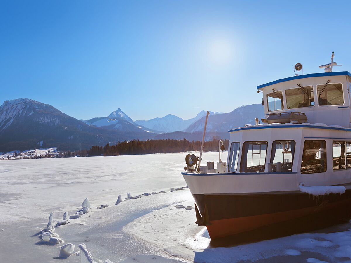 Boot im gefrorenen See mit schneebedeckten Bergen, blauer Himmel und Wintersonne in den Alpen.