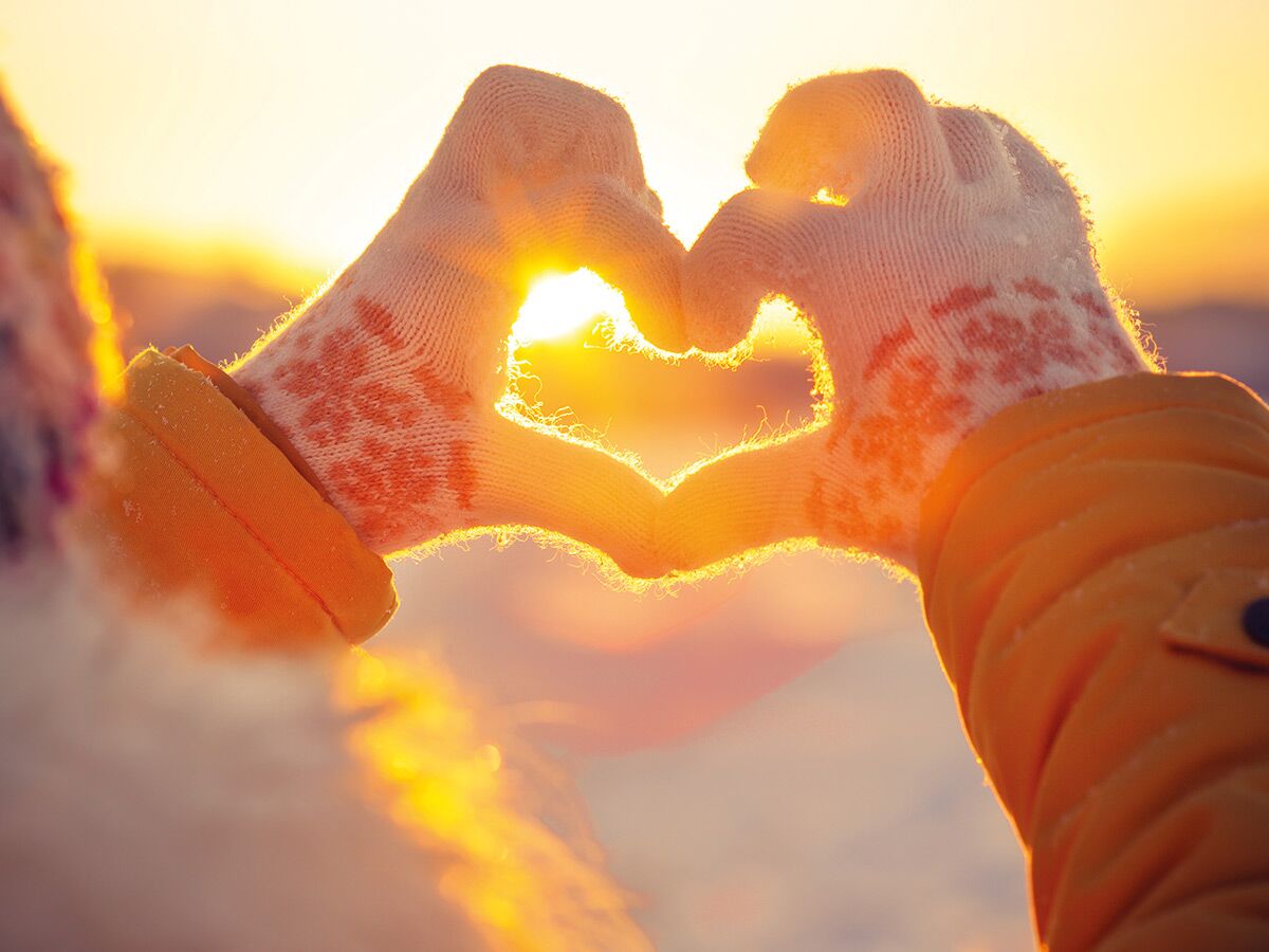 Zwei Hände in Handschuhen bilden ein Herz vor einem warmen, goldenen Sonnenuntergang. Symbolisiert Liebe und Wärme im Winter.