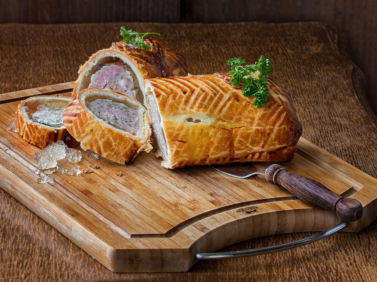 Aufgeschnittene Pâté en Croûte mit Fleischfüllung und Aspik auf einem rustikalen Holzbrett, garniert mit Petersilie.
