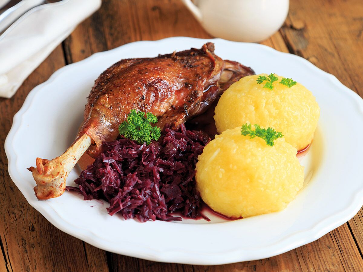 Gebratene Entenkeule mit Rotkohl und Kartoffelklößen auf weißem Teller an Holztisch serviert.