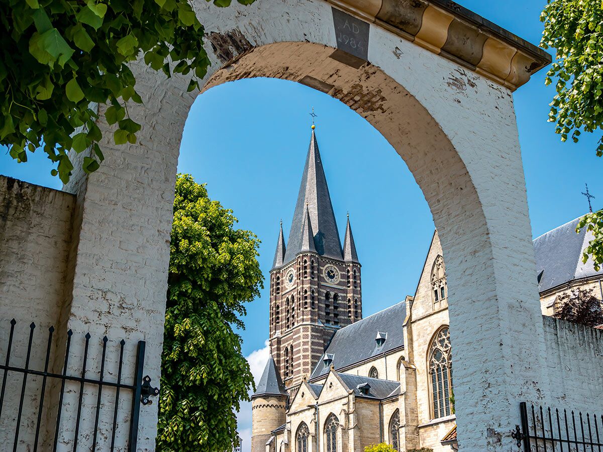 Hoher Kirchturm mit Uhr, gotisches Kirchengebäude, gerahmt von weißem Ziegelbogen mit "AD 1984", umgeben von Grün unter blauem Himmel.