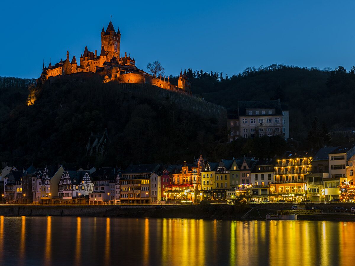 Beleuchtete Reichsburg Cochem bei Nacht über der Mosel mit Stadthäusern und goldenen Lichtreflexionen im Wasser.