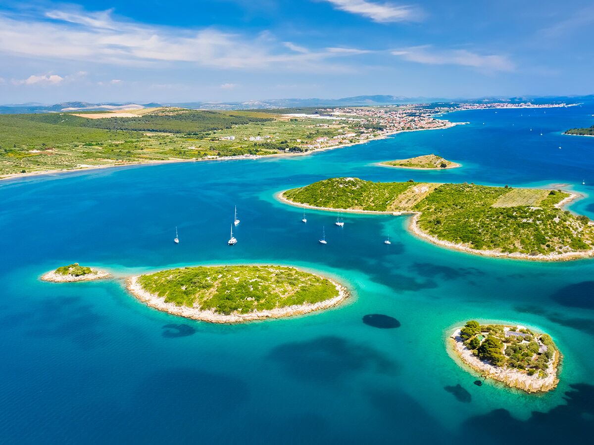 Luftaufnahme der kroatischen Küste mit grünen Inseln, türkisfarbenem Meer, weißen Segelbooten und einer Stadt im Hintergrund.