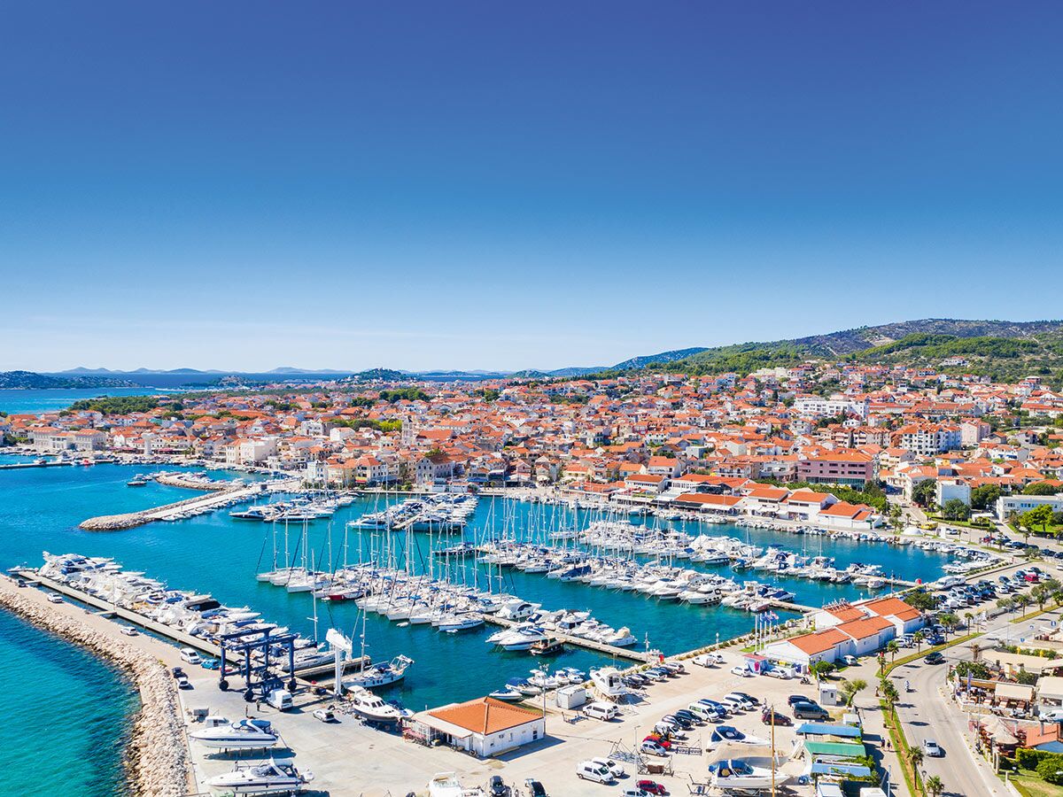 Luftbild einer sonnigen kroatischen Küstenstadt mit lebhaftem Yachthafen voller Segel- und Motorboote und roten Dächern am Hang.