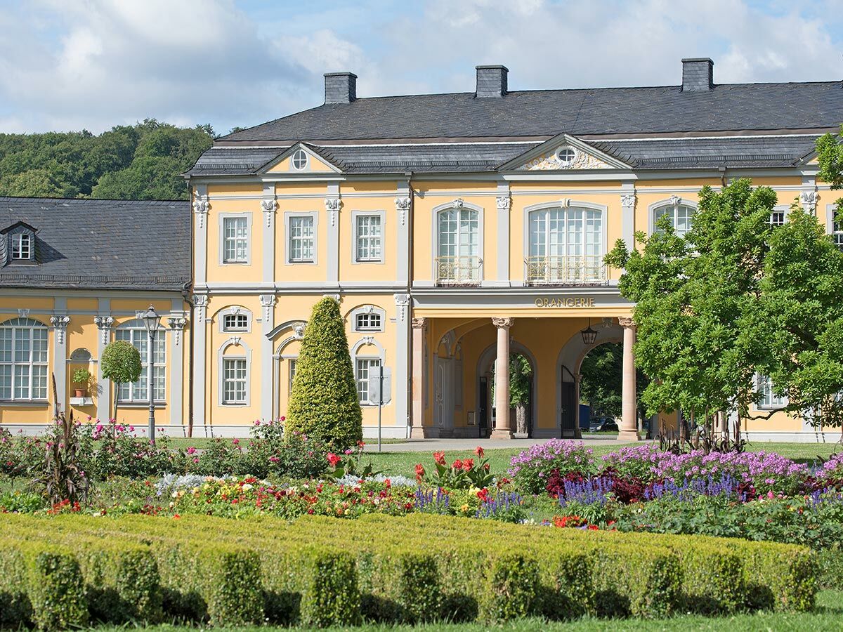 Sonnenbeschienene Orangerie mit gelber Fassade, dunklem Dach, Säulen und Balkonen, umgeben von einem blühenden Barockgarten.