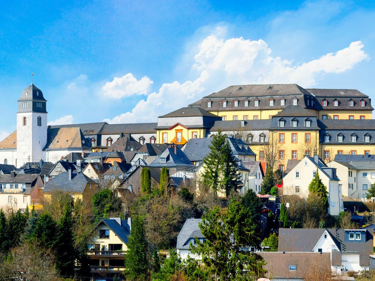 Malerische deutsche Stadtansicht mit weißer Kirche, historischem Gebäude und Häusern auf einem Hügel unter blauem Himmel.