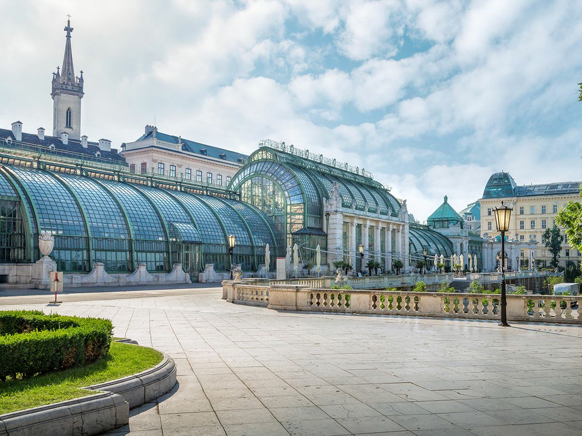 Historisches Palmenhaus Wien im Burggarten mit grüner Jugendstil-Architektur, Park und Kirchturm unter blauem Himmel.