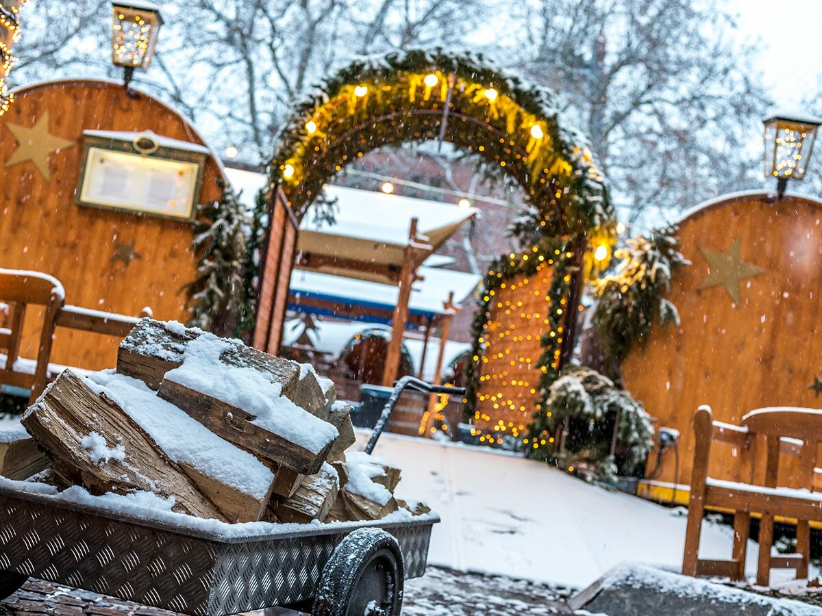 Schneebedecktes Kaminholz in Metallwagen vor festlichem Weihnachtsmarkt mit beleuchteten Holzständen im Schneefall.