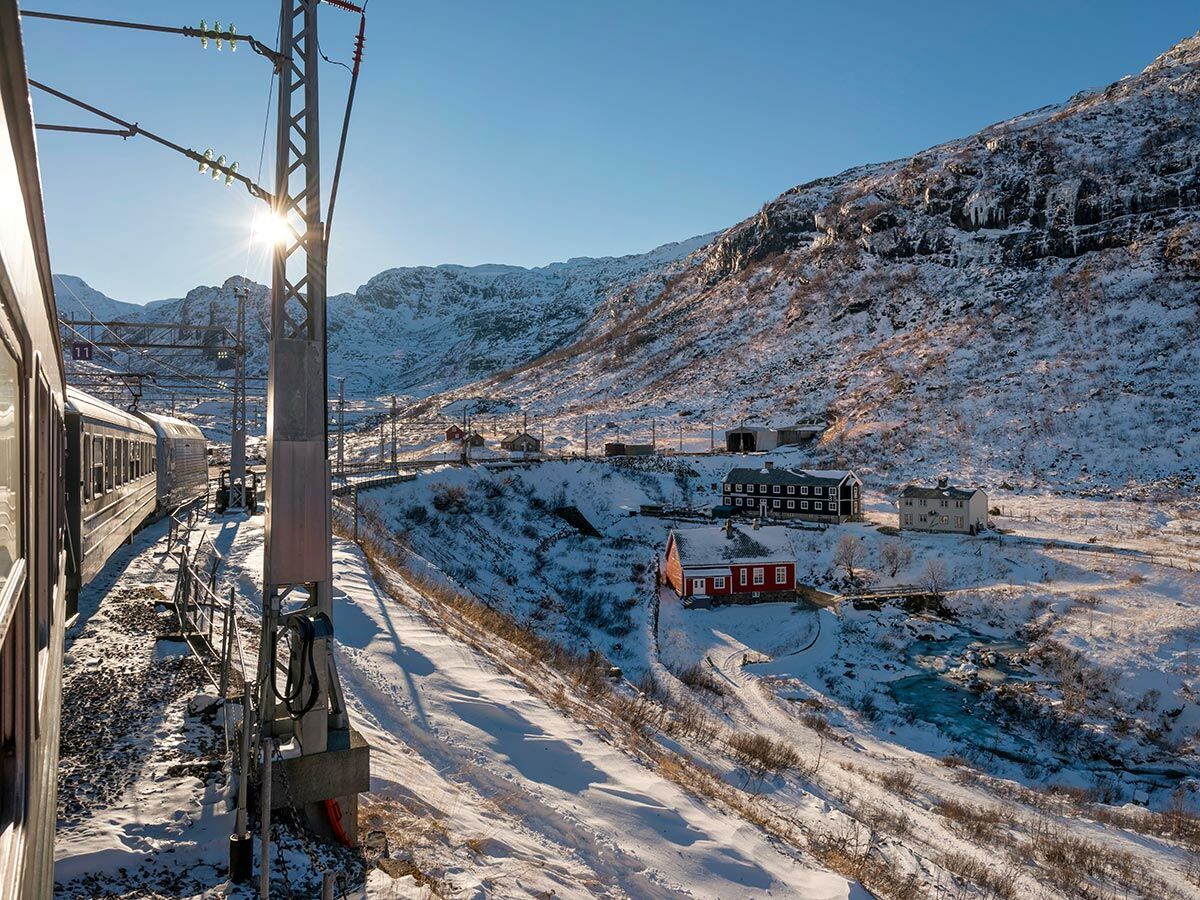 Zug auf der Flåmsbahnstrecke im Winter, umgeben von schneebedeckten Bergen, traditionellen Häusern und eisigem Fluss unter Sonne.