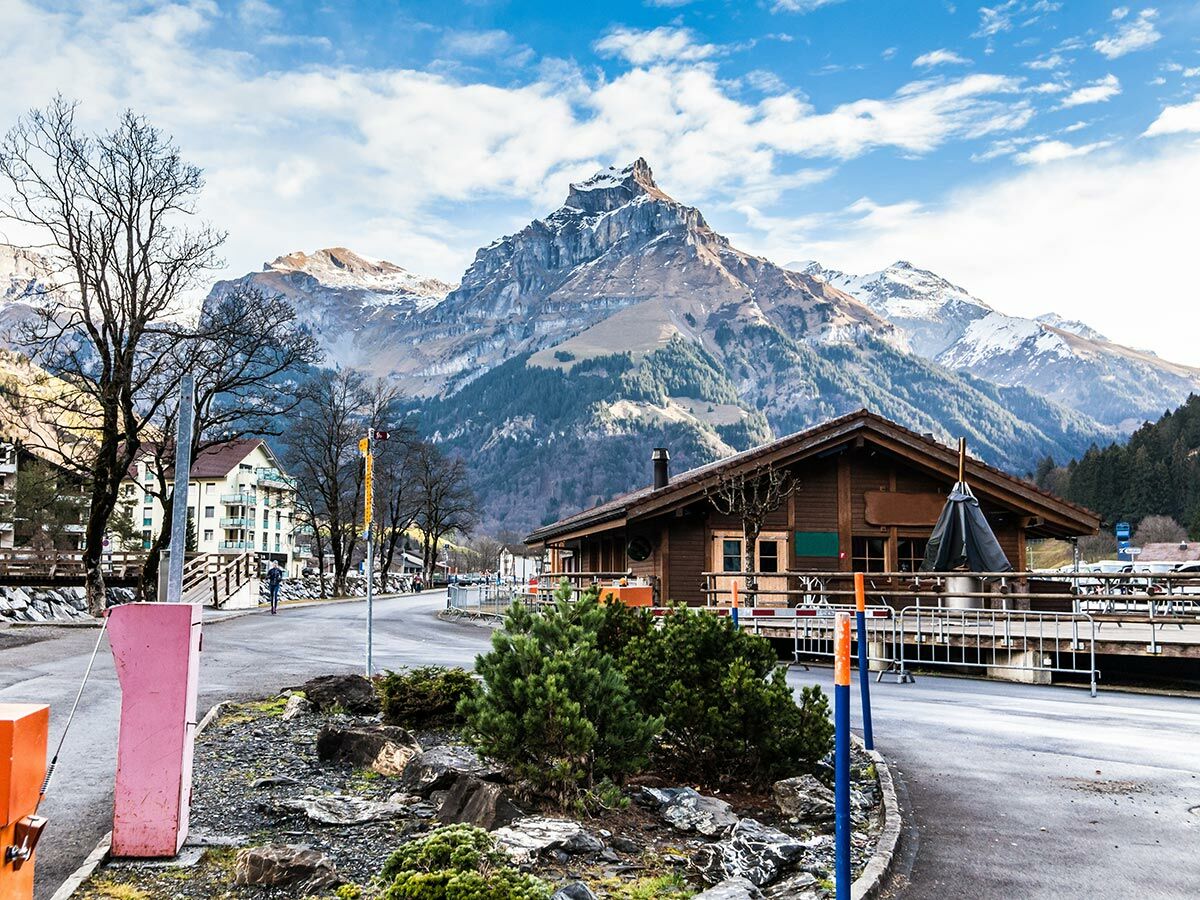 Malerisches Bergdorf mit traditionellem Holzchalet vor majestätischen, schneebedeckten Alpen unter blauem Himmel.