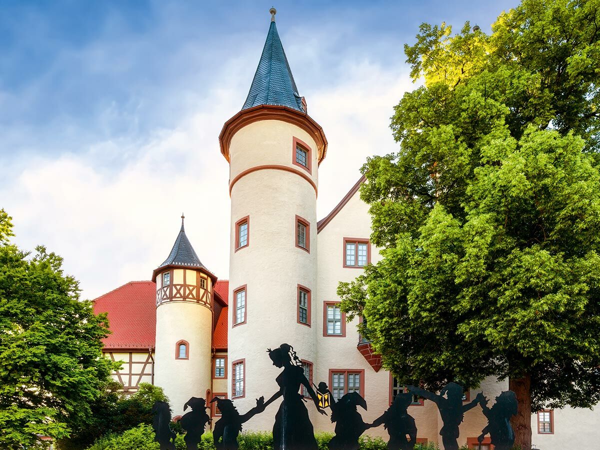 Historisches Märchenschloss mit zwei markanten Türmen, roten Dächern und einer Silhouetten-Skulptur von Sagengestalten im Vordergrund.